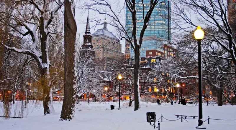 Snow-covered Boston Common at dusk with a gas lamp and a church spire