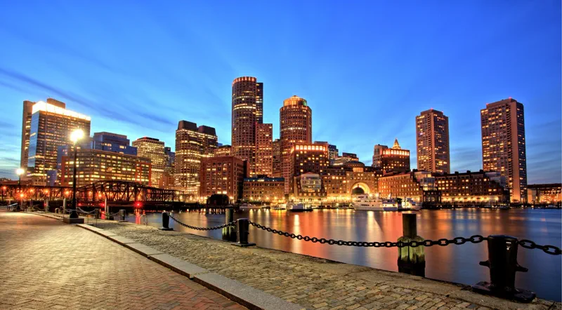 Boston harbor skyline at dusk seen across cobblestones and a chain rail